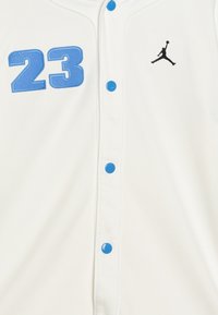Camiseta blanca con el número azul "23" y botones azules. Incluye un logotipo de Jordan en negro. Fabricada con material de malla transpirable.