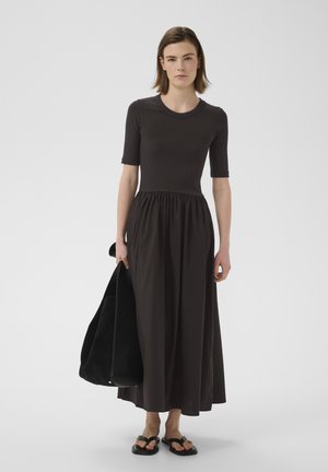 Femme portant un haut noir ajusté à manches courtes avec une longue jupe fluide, portant un grand sac noir et des sandales noires.