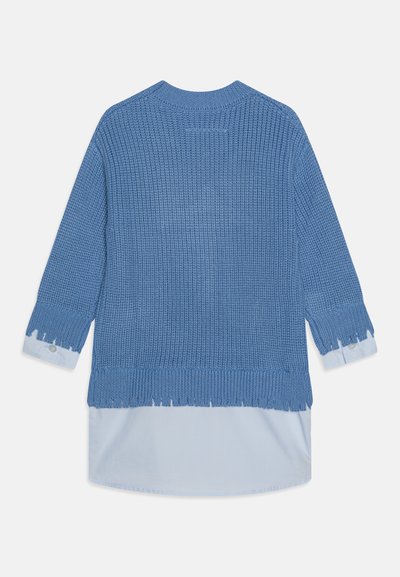 Modrý pletený sveter vrstvený cez svetlomodrú košeľu s dlhými rukávmi a ošúchanými okrajmi na manžetách a na leme.