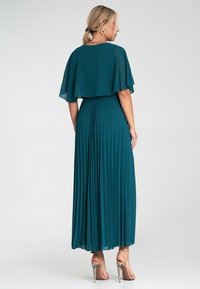 Turkusowa, plisowana sukienka maxi z luźną peleryną, podkreśloną w talii, o półprzezroczystej fakturze, w połączeniu ze srebrnymi sandałami na obcasie.