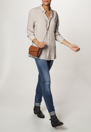 Chemise boutonnée gris clair, jean skinny bleu, bottines noires cloutées aux chevilles et sac bandoulière marron avec une lanière chaîne.