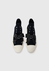 Visoke teniske z navezanim platnenim zgornjim delom v mornarsko modri barvi, belim gumijastim podplatom in velikim črnim usnjenim kiklom čez vezalke, s srebrnimi okovji.