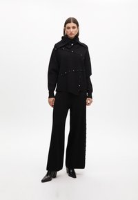 Suéter negro con adornos dorados, cuello oversized y corte asimétrico, combinado con pantalones anchos negros con detalles de botones.