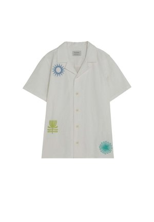 Niño de pie, vistiendo una camisa blanca de manga corta con diseños coloridos de sol, pantalones cortos azul marino y zapatos tipo mocasín azul claro sobre un fondo liso.