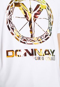 Camisa blanca con un gran gráfico metálico que presenta una figura central, un diseño circular y "DONNAY CARLO COLUCCI" en letras brillantes.