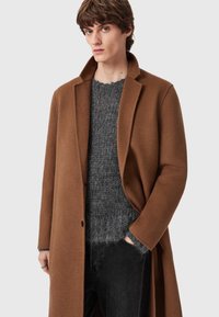 Smeđi vuneni kaput s izrezanim ovratnikom, s jednim dugmetom za zatvaranje. Uparen s teksturiranim sivozim džemperom i crnim hlačama.