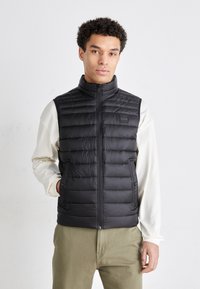 Svart pufferväst med hög krage, dragkedjestängning och två sidofickor, med en logotyp på bröstet och quiltad struktur.