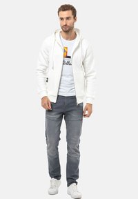 Witte zip-up hoodie met een capuchon met trekkoord, gedragen over een graphic t-shirt. Gepaard met grijze jeans en witte sneakers; casual outfit.
