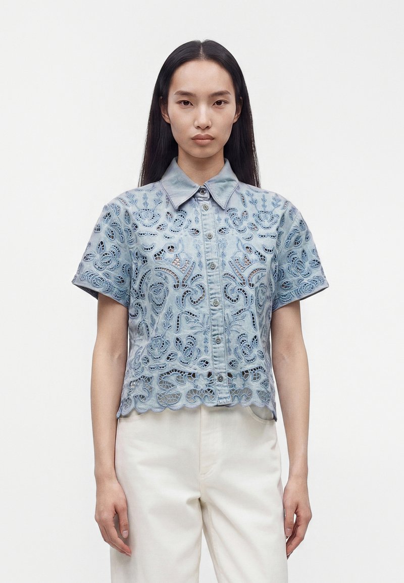 Jeune femme aux longs cheveux noirs porte une chemise en dentelle bleu clair à manches courtes et un pantalon blanc, debout devant un fond clair uni.