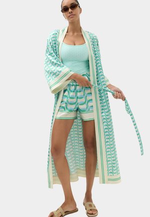 Kvinde iført lyseblå mønstrede shorts, matchende lang kimono, ærmeløs top, solbriller og beige sandaler, der holder kimonoens bælte.