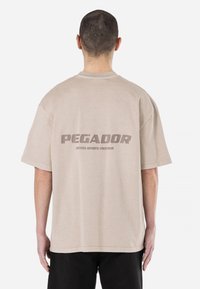 Bež majica oversized kroja s kratkimi rokavi, ki ima na hrbtni strani velik logo "PEGADOR" in besedilo "CERTIFIED AUTHENTIC STREETWEAR".
