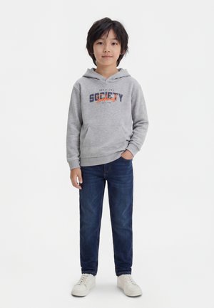 Jeune garçon debout, une main dans la poche, portant un sweat à capuche gris avec un texte, un jean bleu foncé et des baskets blanches sur fond blanc uni.