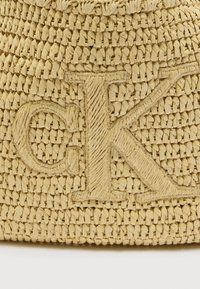 Πλεκτή τσάντα από άχυρο σε ανοιχτό κίτρινο με υφή και σχέδιο. Διαθέτει ευδιάκριτο φυσικό νήμα με το λογότυπο "CK" στο μπροστινό μέρος.