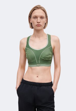 Vrouw draagt een groene sportbh met witte biezen en zwarte sportbroek, staat met handen in de zakken tegen een effen achtergrond.