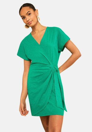 Femme portant une robe portefeuille verte à manches courtes avec un nœud sur le côté et des boucles d'oreilles circulaires dorées, posant avec une main sur la hanche.