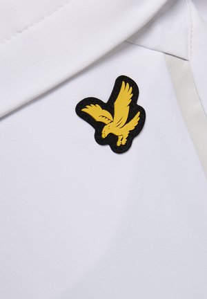 Gelbes Vogel-Emblem mit ausgestreckten Flügeln auf schwarzem Hintergrund, befestigt an einem weißen Hemd mit Kragen.