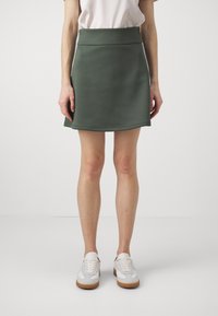 Person som bär en vit topp, olivgrön minikjol och vita sneakers med gummisulor, stående mot en enfärgad ljus bakgrund.