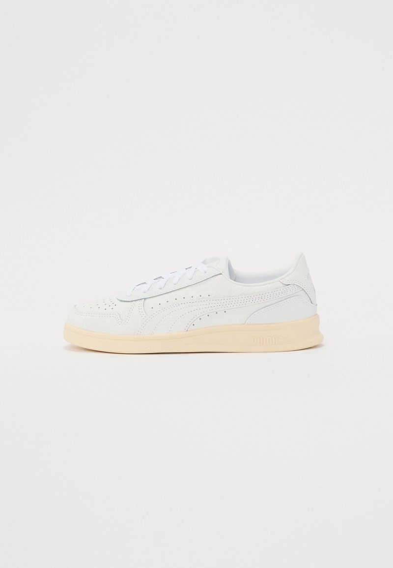 Fehér szintetikus felsőrészes sportcipők, perforált mintázattal, lapos fűzőkkel és krémszínű gumitalppal. A cipő sarokrészén és oldalán logó található.