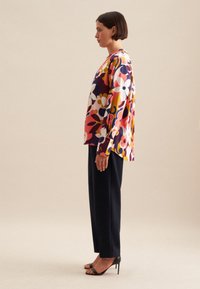 Blusa floreale a maniche lunghe con motivi multicolori nei toni di rosa, giallo e blu navy; abbinata a pantaloni neri e tacchi a fascia.