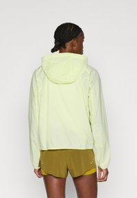 Ελαφρύ, φλις μπουφάν με κουκούλα σε φωτεινό κίτρινο χρώμα με λεία υφή, διαθέτοντας ελαστικές μανσέτες και διακριτικό λογότυπο κοντά στο τελείωμα. Συνδυάζεται με χακί σορτς.