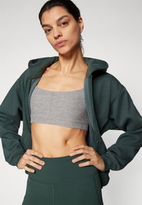 Grønn zip-up hettegenser med en myk tekstur, grå croppet tank topp under, matchende høytlivede grønne treningsbukser som viser en atletisk passform.