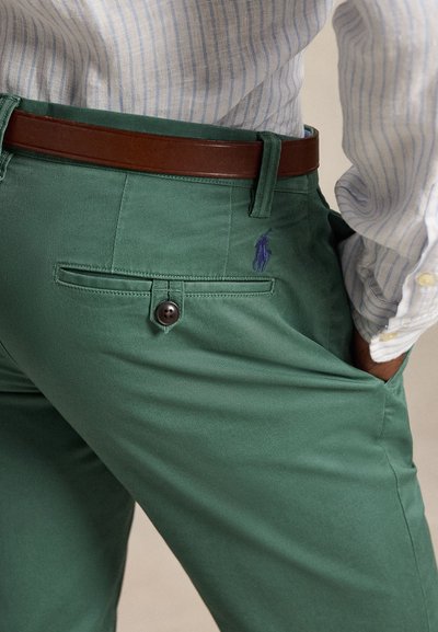 Mand iført grønne bukser med et brunt bælte og en hvid og blåstribet skjorte stukket ned, hånd i lommen.