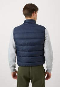 Navyblaue Steppweste mit horizontaler Steppung, hohem Kragen und glatter Textur, getragen über einem hellgrauen Langarmshirt.