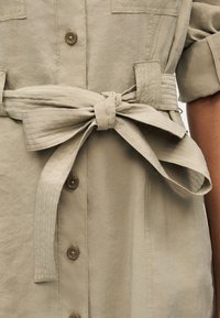 Μπεζ ένδυμα με κουμπιά και φαρδιά ζώνη από ύφασμα δεμένη σε φιόγκο στη μέση, με το μανίκι στο δεξί χέρι ανασηκωμένο.