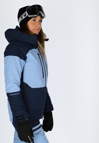 Blue and navy ski jacket with a hood and zip pocket, paired with black gloves. Features contrast color blocking and logo accents.
