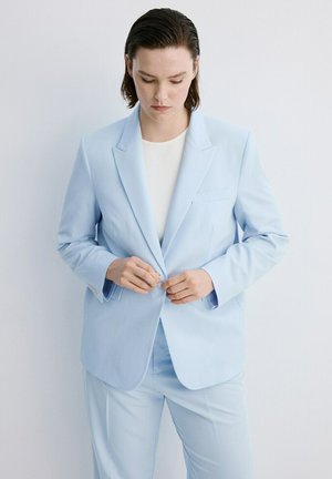 Vrouw met kort donker haar die een lichtblauwe colbert met bijpassende broek en witte top dichtknopt, staand tegen een witte achtergrond.