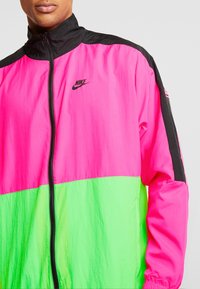 En ljus rosa och grön Nike vindjacka med svart dragkedja och svarta detaljer, bärs av en person viss synlig från halsen till midjan.