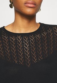 Donna che indossa orecchini ovali d'oro e un top in maglia nera con una scollatura a yoke trasparente a zigzag e scollatura rotonda.
