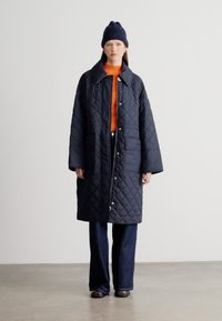 Μακρύ μπουφάν σε στρώσεις σε ναυτικό μπλε χρώμα με γιακά, κούμπωμα με κουμπιά πατιλέτα και δύο μεγάλες μπροστινές τσέπες, φορεμένο πάνω από πορτοκαλί πουλόβερ και σκούρο παντελόνι.