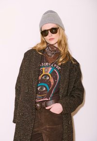 Femme portant un bonnet gris, des lunettes de soleil sombres, un foulard en bandana marron à motifs, un t-shirt à motif, un manteau texturé et un pantalon marron, avec une main dans la poche.