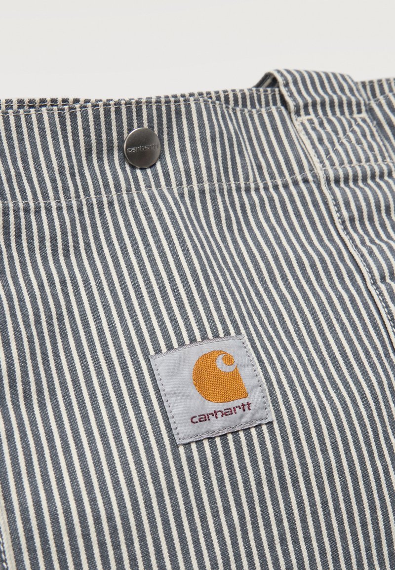 Gros plan sur un tissu rayé bleu et blanc avec un patch logo Carhartt cousu et un bouton en métal sur la ceinture d'un vêtement.