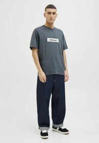 Grå bomulds T-shirt med et rektangulært hvidt og sort grafisk mærke. Båret med løstsiddende mørkeblå jeans og sorte sneakers.
