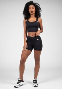 Zwarte sportbeha met mesh-details, gecombineerd met hoge zwarte shorts. Inclusief witte logo-accenten. Model draagt zwarte sportschoenen.