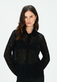 Blusa in pizzo nero con maniche lunghe. Presenta un colletto e chiusura frontale con bottoni. Il motivo in pizzo intricato aggiunge texture al capo.