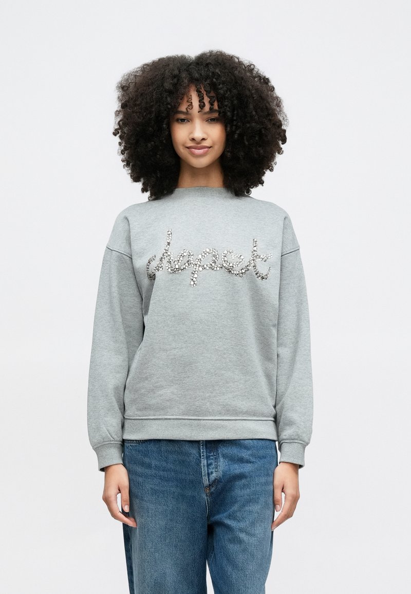 Pelēks džemperis ar apaļu kaklu, rotāts ar mirdzošu uzrakstu "respect" no rhinestones. Kombinēts ar ikdienišķiem zilajiem džinsiem.