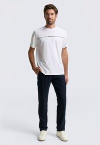 Wit T-shirt met korte mouwen, zwart logo en lijndetail, gecombineerd met marineblauwe cargobroek en witte sneakers. Simpele en casual outfit.