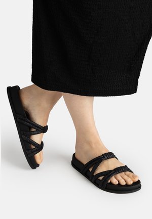 Feet wearing black strappy flat sandals with textured fabric straps, shown with cropped black pants against a plain background.