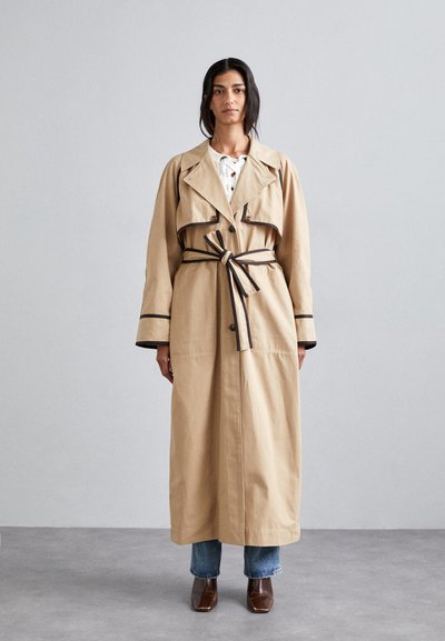 Manteau beige avec une taille ceinturée, des bordures sombres contrastantes, des manches longues et une fermeture à boutons devant, superposé sur un jean bleu et des chaussures marron.