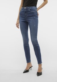 Uzane traperice u tamnoplavoj boji, visoki struk, s istrošenim uzorkom i pet džepova; uparene s crnim cipelama na petu.