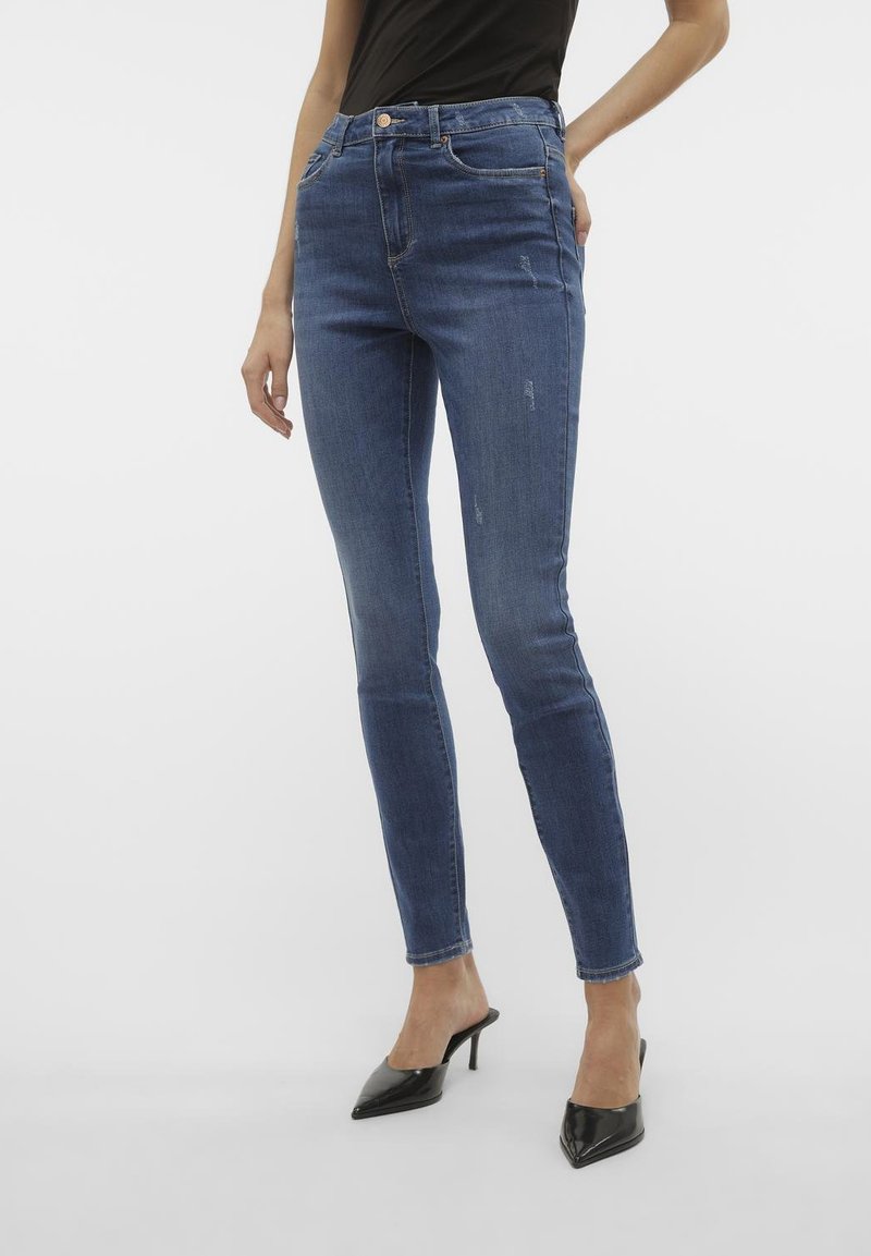 Uzane traperice u tamnoplavoj boji, visoki struk, s istrošenim uzorkom i pet džepova; uparene s crnim cipelama na petu.