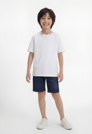 Ragazzo sorridente in piedi, che indossa una maglietta bianca, pantaloncini di jeans scuri e scarpe da ginnastica bianche su uno sfondo bianco neutro.