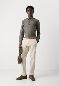 Männeroutfit mit einem dunkelgrünen Hemd, beigefarbenen Hosen und braunen Sneakern, accessoriert mit einer grünen Umhängetasche.