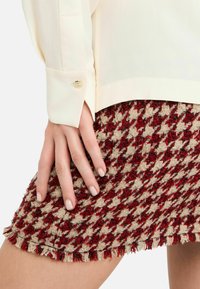 Blusa beige chiaro con dettagli di bottoni dorati, abbinata a una gonna a quadretti houndstooth rossa e beige con orlo frangiato.