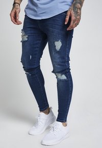 Jeans skinny de color azul oscuro, rasgados y con detalles deshilachados, fabricados en denim, combinados con zapatillas blancas. Tejido suave con detalles desgastados.