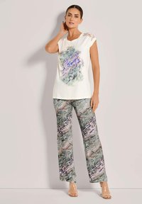 Vrouw die staat in een witte top met korte mouwen met multicolor abstract print, gecombineerd met brede broek in een bijpassend abstract patroon en transparante hakken.
