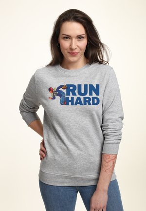 Femme portant un sweat-shirt gris avec un joueur de football vintage et un texte bleu vif "COURIR DUR", debout avec une main sur la hanche, fond clair.